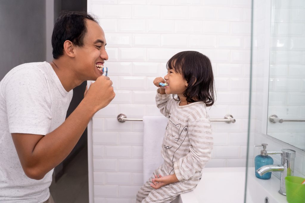 Child with dad learns how to brush their own teeth on a daily routine before bed.